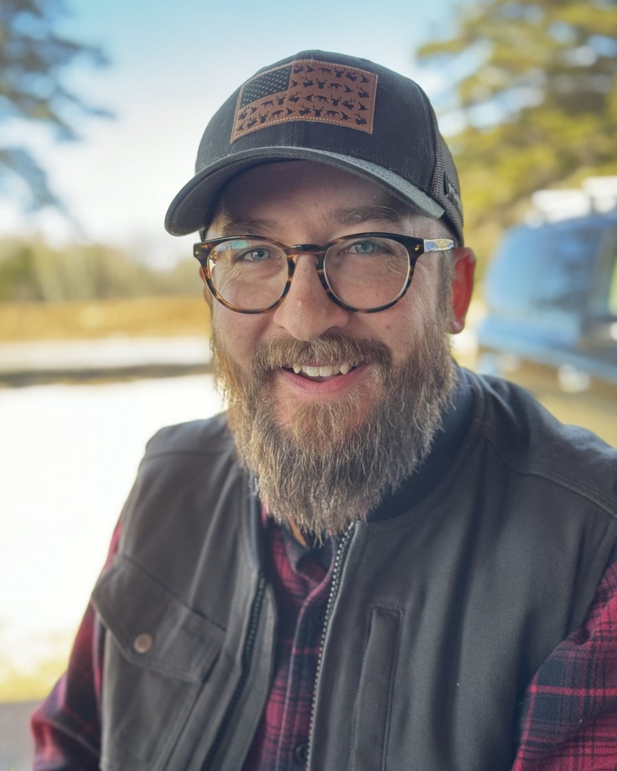 Rob Rebhan, smiling, in a trucker cap, glasses, and a flannel shirt outdoors